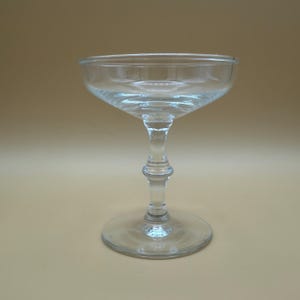 May include: A clear, stemmed champagne coupe glass. The glass features a wide, shallow bowl, a slender stem with a decorative bulbous section, and a circular base. The glass is transparent, allowing light to pass through, and is set against a neutral background.