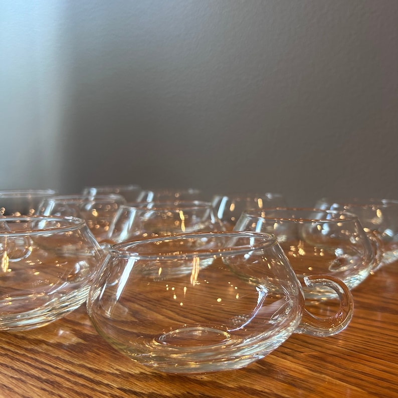 May include: A group of clear glass cups with rounded bottoms and handles. The cups are arranged in a staggered pattern on a wooden surface.