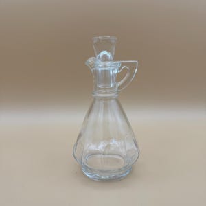 May include: A clear glass cruet with a wide, tapered base and a narrow neck. The vessel features a small integrated handle on the side, a pouring spout, and a removable glass stopper that sits securely within the opening at the top of the neck.