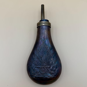 May include: Antique powder flask with a pear-shaped body and a long spout. The flask has a dark, iridescent finish with an embossed design featuring crossed flags, cannons, and other military motifs. The spout is made of metal.