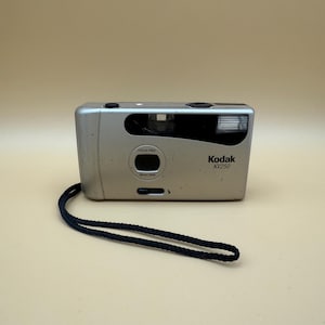 Puede incluir: Cámara de película Kodak KV250 de 35 mm plateada con correa negra. La cámara tiene una lente sin enfoque y un flash incorporado. El logotipo de Kodak y el número de modelo están impresos en la parte frontal.