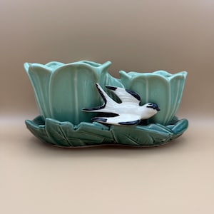 May include: A ceramic planter set featuring two light green tulip-shaped vessels attached to a dark green leaf-shaped base. A white and black swallow bird figure is attached to the front of the vessels, creating a cohesive decorative piece.