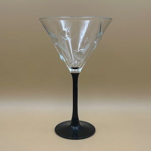 Könnte beinhalten: Ein klares Martiniglas mit schwarzem Stiel und Fuß. Das Glas hat ein geätztes Kirschdesign auf der Schale. Der Stiel ist lang und schlank, und der Fuß ist rund. Das Glas ist leer.