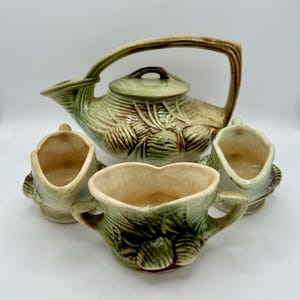 Peut inclure: Service à thé en céramique vintage avec une théière et trois crémier. L'ensemble présente un motif texturé vert et marron, avec une grande anse sur la théière. Les crémier ont un design en forme de cœur.