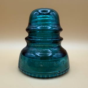 Könnte beinhalten: Ein vintage elektrischer Isolator aus blaugrünem Glas mit abgerundeter Oberseite und abgestuften Rillen. Die Basis zeigt den geprägten Text HEMINGRAY. Dieses schwere, durchscheinende Objekt hat eine glatte, geformte Oberfläche mit feinen Blasen.