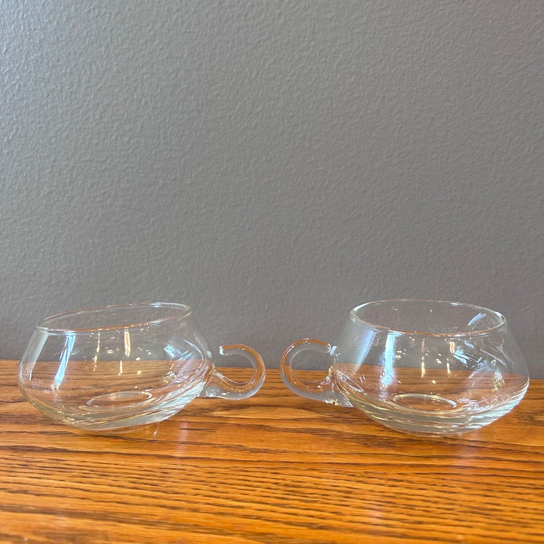 May include: Two clear glass cups with rounded bottoms and handles. The cups are sitting on a wooden surface.