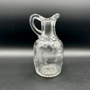 May include: A clear glass pitcher with a handle and a spout. The pitcher has a rounded body and a flared neck. Delicate leaf and vine etchings decorate the body. The pitcher is empty and sits on a dark surface.