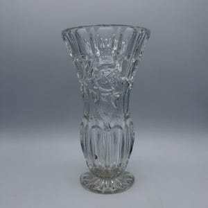 May include: Clear glass vase with a flared top and a round base. The vase features a detailed floral and leaf pattern, with vertical ribbed detailing along the body. The vase is designed to hold flowers and other decorative items.