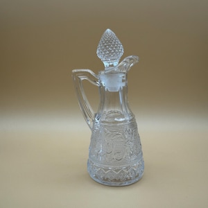 Peut inclure: Une burette en verre transparent présentant un motif en relief complexe sur le corps, une anse robuste et un bouchon en verre assorti avec un sommet texturé et pointu. Le récipient a une base large qui s'affine vers le col et un petit bec verseur.