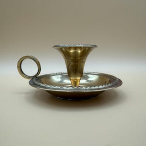 Könnte beinhalten: Ein Vintage-Kerzenhalter aus Messing und Silber. Der Kerzenhalter hat eine ausgestellte Oberseite und einen kreisförmigen Sockel mit dekorativem Rand. Ein kleiner Griff ist an der Seite des Sockels angebracht. Der Kerzenhalter ist etwa 10 cm hoch.