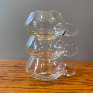 May include: Three clear glass cups with handles stacked on top of each other. The cups are round and have a slight curve to them.