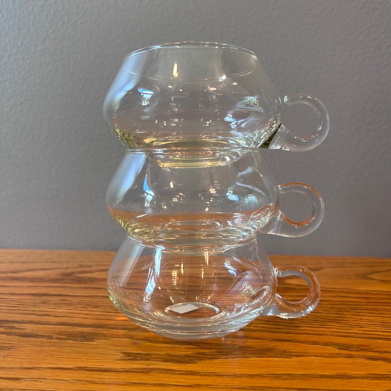 May include: Three clear glass cups with handles stacked on top of each other. The cups are round and have a slight curve to them.
