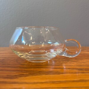 May include: A clear glass cup with a rounded bottom and a single handle. The cup is sitting on a wooden surface.