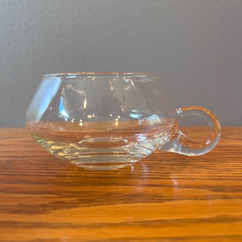 May include: A clear glass cup with a rounded bottom and a single handle. The cup is sitting on a wooden surface.