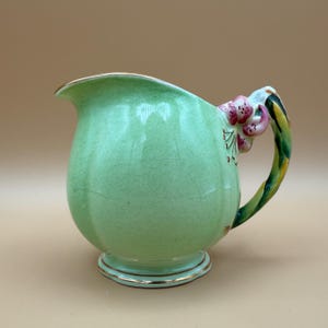 May include: A vintage green ceramic jug with a gold rim and base. The handle is a twisted design with green, yellow, and gold colours. Pink and red floral accents adorn the jug's handle. The jug has a bulbous shape.
