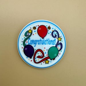 Puede incluir: Una etiqueta de regalo redonda y blanca con un borde azul. Presenta un diseño de globos rojos, verdes y morados con la palabra "Congratulations!" en azul. Estrellas amarillas y puntos de confeti completan el aspecto festivo.