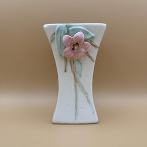 May include: A white ceramic vase with a flared top and base, featuring a raised, three-dimensional pink flower with a brown center. The design includes painted light green leaves and thin brown stems extending downward along the front surface.