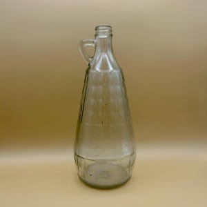 May include: A clear glass jug with a handle. The jug has a wide, ribbed body that tapers to a narrow neck and a small opening. The base of the jug has a textured design. The jug is empty and sits against a light brown background.
