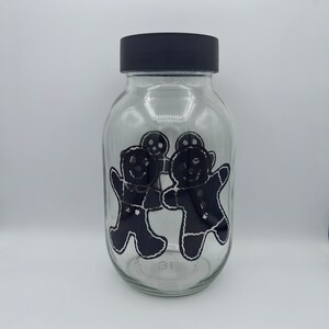 Könnte beinhalten: Ein klares Glas mit einem schwarzen Deckel. Das Glas hat ein schwarz-weißes Lebkuchenmann-Design. Das Glas ist zylindrisch und hat eine weite Öffnung. Das Design umfasst vier Lebkuchenmänner.