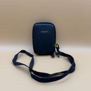 May include: A black, rectangular Ruggard camera case with a black strap. The case has the word "Ruggard" printed on the front. The strap is attached to the case with a black clip.