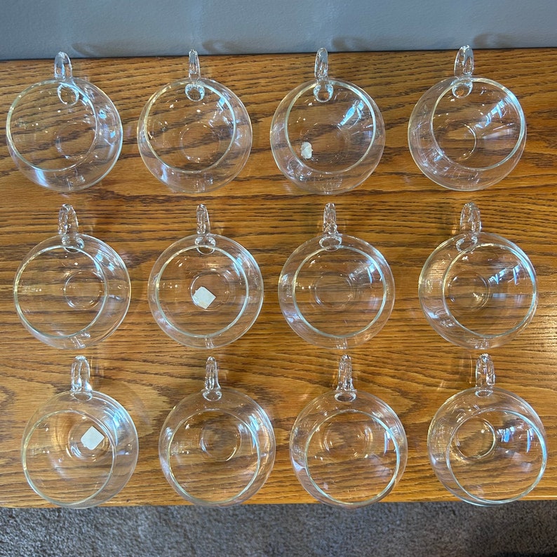 May include: Twelve clear glass teacups with handles, arranged in three rows of four on a wooden surface.