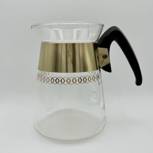 Puede incluir: Una jarra de vidrio vintage con una banda de metal dorada y un asa negra. La jarra tiene un cuerpo de vidrio transparente con un patrón dorado decorativo alrededor del centro. El asa es curva y está unida a la jarra con una pieza de metal dorada.