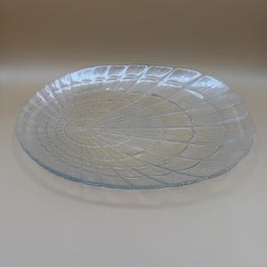 May include: A clear, oval-shaped glass serving platter with a textured, radial design. The platter has a slightly raised edge and a diameter of approximately 12 inches. The design resembles a stylized flower or shell.