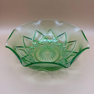 Puede incluir: Un cuenco de vidrio verde claro con un borde festoneado. El cuenco presenta un diseño floral en el centro y en los lados. El cuenco es de vidrio transparente y tiene una estética vintage.