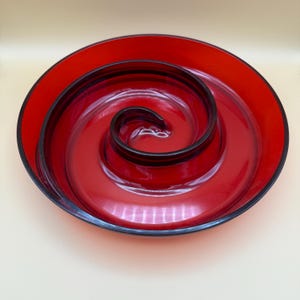 May include: A vibrant red glass serving dish with a unique spiral design. The dish features a deep red color with a black trim around the edges and the spiral center. The spiral design adds a modern touch to this decorative piece.