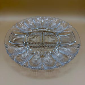 May include: A clear, oval-shaped glass serving dish with multiple compartments. The dish features a detailed, textured design with a central section and surrounding compartments, ideal for serving appetizers or snacks. The edges have a decorative pattern.