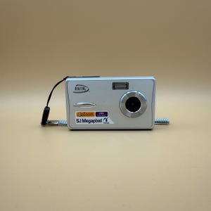 May include: Silver digital camera with a lens, flash, and wrist strap. The camera has a 4x zoom and 5.1-megapixel resolution. The word "DIGITAL" is printed on the top of the camera.