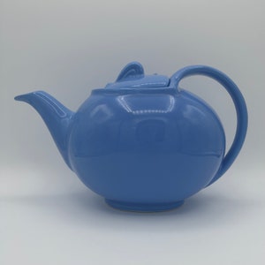 May include: A light blue ceramic teapot with a rounded body and a curved spout. The teapot has a lid with a small handle.