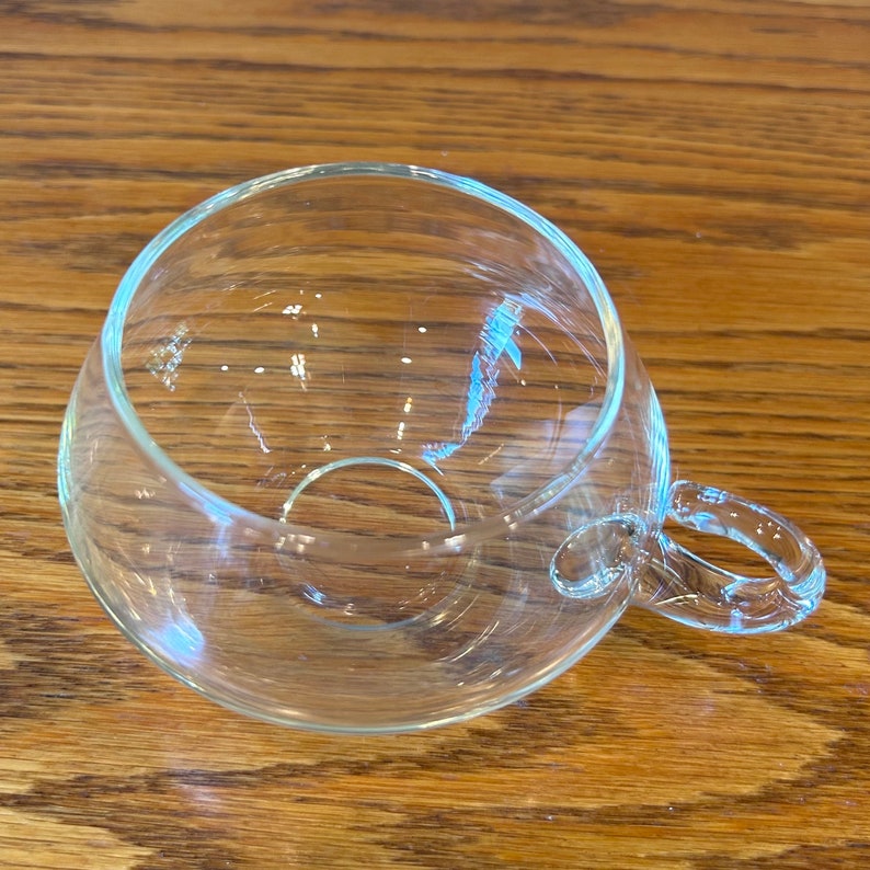 May include: A clear glass cup with a rounded bottom and a single handle. The cup is empty and sitting on a wooden surface.