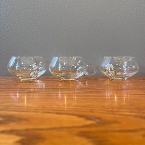 May include: Three clear glass cups with a rounded bowl shape and a single loop handle. The cups are sitting on a wooden surface.