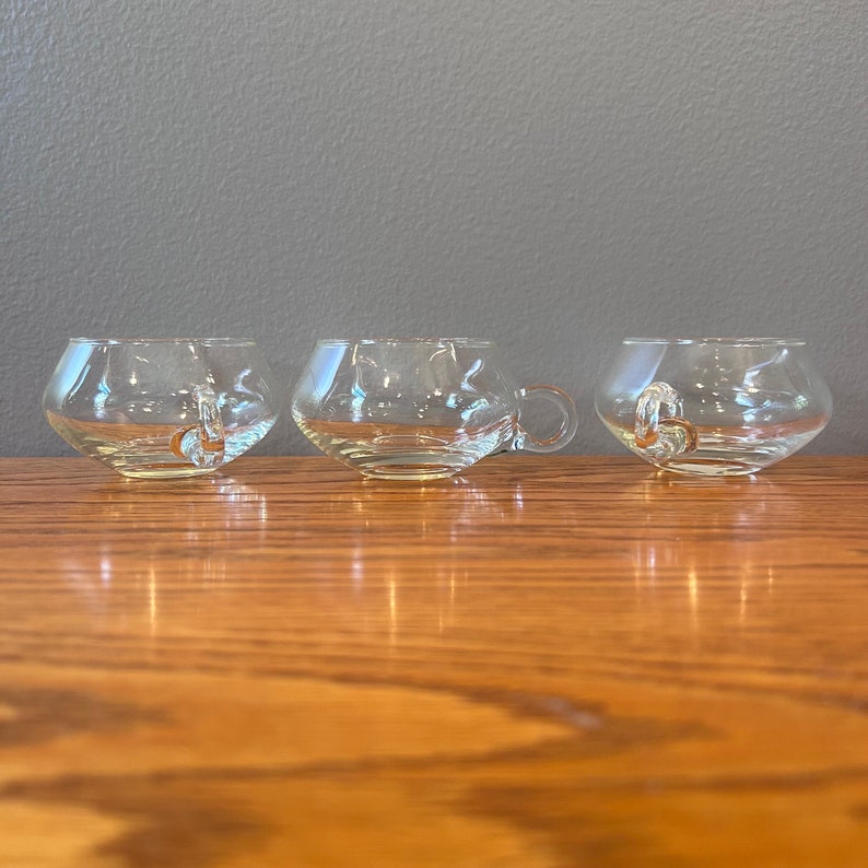 May include: Three clear glass cups with a rounded bowl shape and a single loop handle. The cups are sitting on a wooden surface.