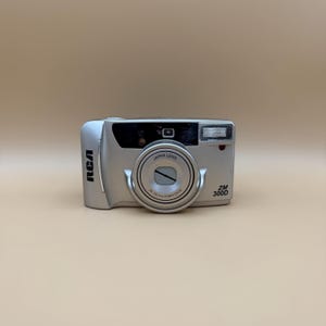 May include: Silver RCA ZM 300D digital camera with a Japan lens. The camera has a flash, a lens, and the RCA logo on the side. The camera is on a beige background.