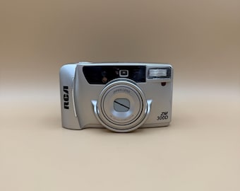 Fotocamera a pellicola RCA ZM 300D con zoom motorizzato 30-120 mm, colore oro con dettagli argento e neri, stile classico "punta e scatta", pezzo da collezione per gli appassionati di fotografia.
