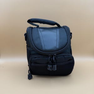 May include: Black camera bag with a top handle and a shoulder strap attachment. The bag has a zippered main compartment and a front pocket. The exterior is made of a textured, durable material. The bag measures approximately 8 inches in height.