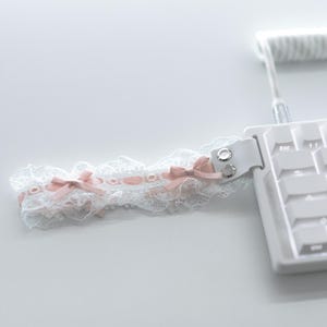 Pode incluir: Um teclado mecânico branco com um apoio de pulso decorativo de renda. O apoio de pulso é branco com laços e detalhes rosa. Um cabo branco enrolado está conectado ao teclado.
