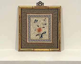 May include: A framed floral embroidery with a gold bamboo-style frame. The embroidery features a central orange flower, a butterfly, and delicate stems. The frame has a decorative top piece and a patterned fabric backing.
