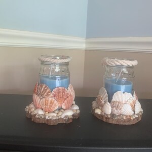 May include: Two glass jar candles with blue wax, each featuring a thick rope trim around the rim and a base decorated with an assortment of layered, light brown and white seashells attached to a circular wooden slice.