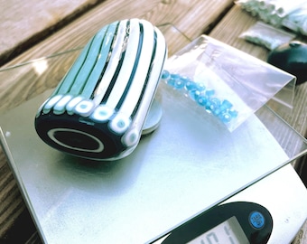 May include: A glass object with vertical teal and white stripes resting on a digital scale that displays a weight of 1.01 ounces. Next to the object is a small clear plastic bag containing several tiny light blue beads with white centers.