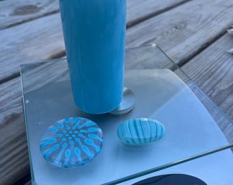 May include: A cylindrical light blue glass object stands on a digital scale next to two smaller glass pieces. One piece is a flat circular disc with a blue and white dot pattern, and the other is an oval-shaped glass stone with blue and white stripes.