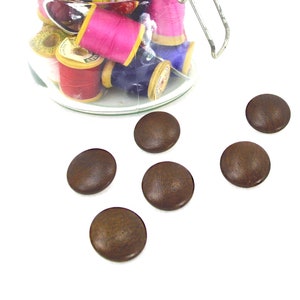 May include: A glass jar filled with colorful spools of thread and several brown, round buttons. The buttons are scattered on a white surface. The thread colors include pink, red, yellow, and blue.