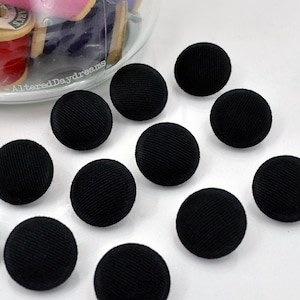 May include: A collection of round, black fabric-covered buttons with a visible ribbed texture. The buttons are arranged in a scattered pattern. A clear glass container holding colorful spools of thread with the text AlteredDaydreams is visible.