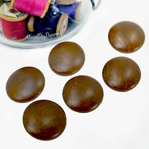May include: Six round, brown, leather-covered buttons with a smooth, slightly domed surface. In the background, a clear glass container holds several wooden spools of thread in various colors, including pink and blue, with the text AlteredDayDreams.