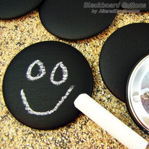 May include: Black chalkboard buttons with a smiley face drawn on one button using white chalk. A piece of white chalk is lying next to the buttons.