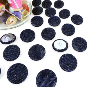 May include: A collection of navy blue denim-covered buttons scattered on a white surface. The buttons are round with metal backs. A glass jar filled with spools of colorful thread is visible in the background, suggesting a sewing or craft theme.