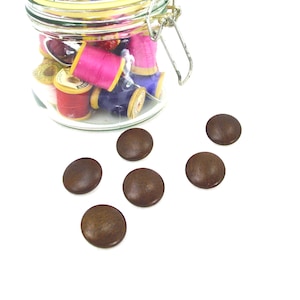 May include: A glass jar filled with spools of colorful thread, including pink, red, and blue. Scattered around the jar are several brown, round buttons. The image is set against a white background, highlighting the sewing supplies.