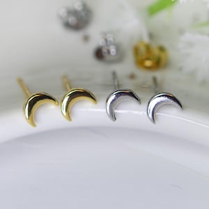 May include: A collection of crescent moon stud earrings in gold and silver tones. The earrings are displayed on a white, star-speckled surface, with white flowers and green stems in the background. The earrings are small and delicate, with a simple, minimalist design.